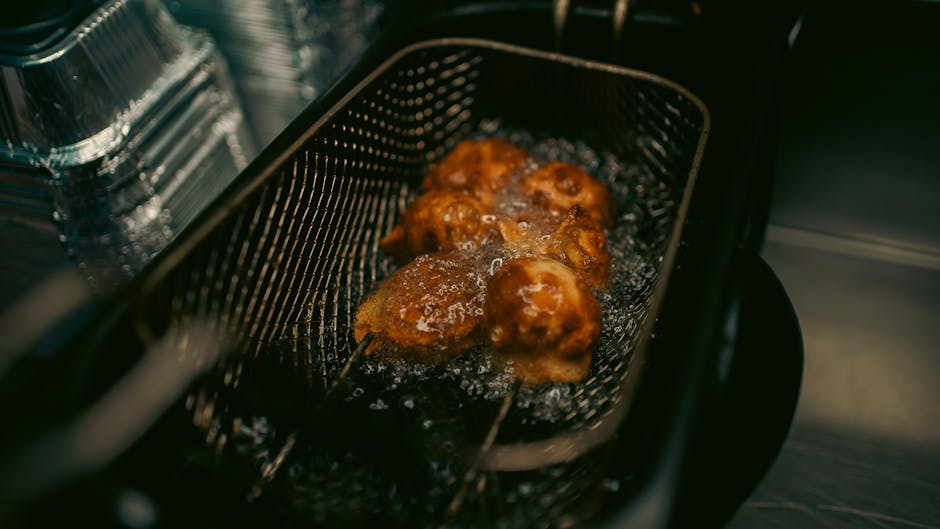 Close-up of four pieces of breaded food, likely chicken nuggets or similar, frying in a black wire basket immersed in hot oil within a deep fryer. The food appears golden-brown and crispy, with some bubbling oil surrounding them, indicating they are being deep-fried. The fryer’s interior features a stainless steel side panel, and a clear view of the bubbling oil and the textured basket. The scene is well-lit, emphasizing the crispy surface of the fried items, highlighting the cleaning and maintenance aspects associated with oven and kitchen appliance surface cleaning, as promoted by Oven Cleaning Merton on their page about grease disposal and deep cleaning.