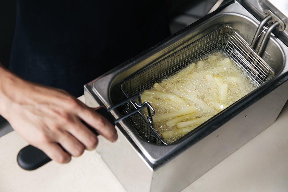 A person is removing a wire basket filled with multiple potato fries from a deep fryer with bubbling hot oil. The deep fryer has a stainless steel exterior and is positioned on a white countertop in a kitchen setting with bright lighting. The person's hand is grasping the basket's handle, and their arm is partially visible, indicating an active deep cleaning or food preparation process. The surrounding area appears clean and well-maintained, reflecting hygienic standards for domestic or commercial food surface cleaning, consistent with the services offered by Oven Cleaning Merton. The scene emphasizes surface cleaning and deep cleaning for food safety and hygiene in accordance with Merton Council guidelines for grease disposal as highlighted on the page about rules for grease disposal.