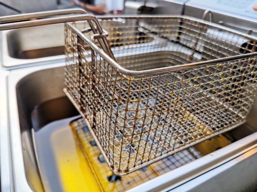 Close-up of four pieces of breaded food, likely chicken nuggets or similar, frying in a black wire basket immersed in hot oil within a deep fryer. The food appears golden-brown and crispy, with some bubbling oil surrounding them, indicating they are being deep-fried. The fryer’s interior features a stainless steel side panel, and a clear view of the bubbling oil and the textured basket. The scene is well-lit, emphasizing the crispy surface of the fried items, highlighting the cleaning and maintenance aspects associated with oven and kitchen appliance surface cleaning, as promoted by Oven Cleaning Merton on their page about grease disposal and deep cleaning.