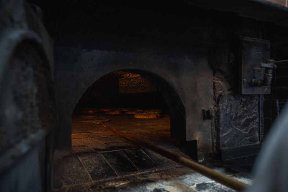 Close-up of a commercial bakery oven with a dark, soot-covered exterior and a glowing interior revealing freshly baked pizzas on the oven floor. The oven's stainless steel and brick surfaces show signs of extensive use, and a long pizza peel is partially visible inside. The surrounding area appears clean and well-maintained, emphasizing thorough surface cleaning and sanitisation by Oven Cleaning Merton. Bright ambient lighting highlights the oven's condition, indicating a professional deep cleaning process suitable for bakery and food service environments.