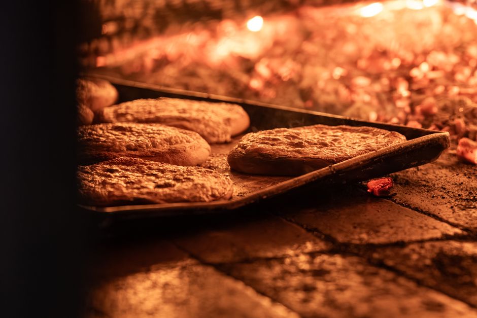 Photograph taken from outside a bakery oven showing several freshly baked bread rolls on a metal tray inside the glowing oven; the brick interior of the oven is visible with a warm orange hue from the heating elements, and the surface of the dough appears golden brown. The tray and bread are positioned towards the front of the oven, with the brick floor stained from baking. The lighting highlights the texture of the baked goods and the oven's interior, reflecting a clean and properly maintained environment, showcasing professional surface cleaning and deep cleaning practices by Oven Cleaning Merton in preparation for domestic or commercial baking needs.