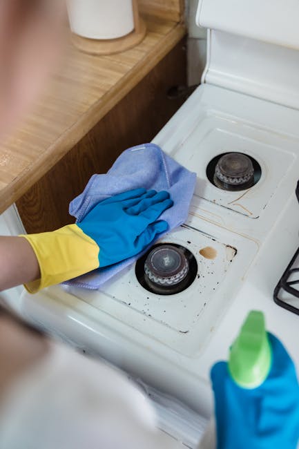 A person wearing blue and yellow rubber gloves is cleaning a glass oven door with a blue cloth, removing dirt and grease to achieve a spotless surface. The oven's interior is visible through the glass