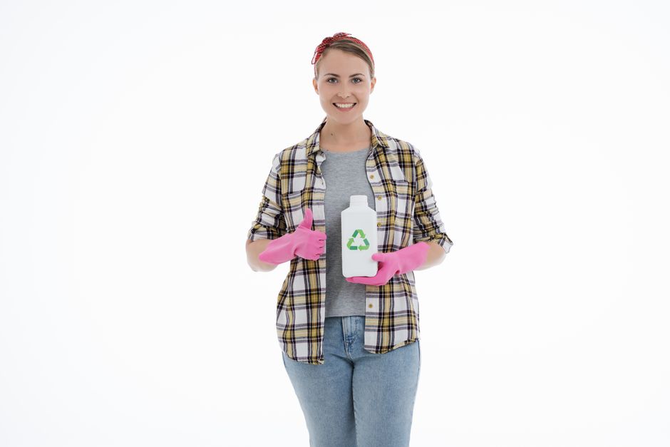 A young woman with a friendly smile stands in a well-lit, white room dressed in casual clothing including a plaid shirt, jeans, and pink rubber cleaning gloves. She is holding a white recycling-compat