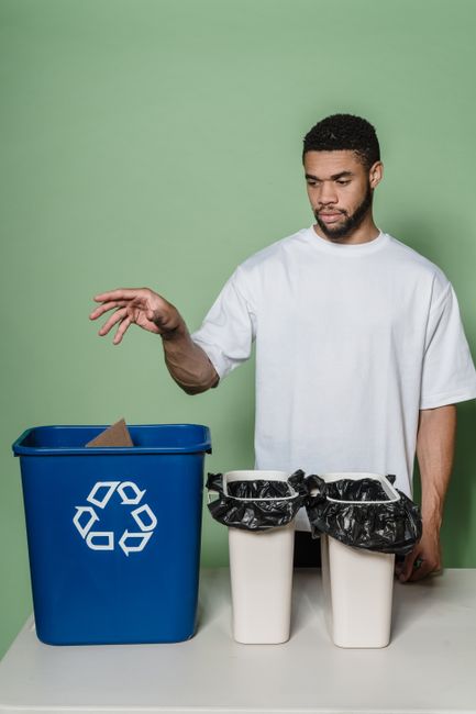 A young woman with a friendly smile stands in a well-lit, white room dressed in casual clothing including a plaid shirt, jeans, and pink rubber cleaning gloves. She is holding a white recycling-compat
