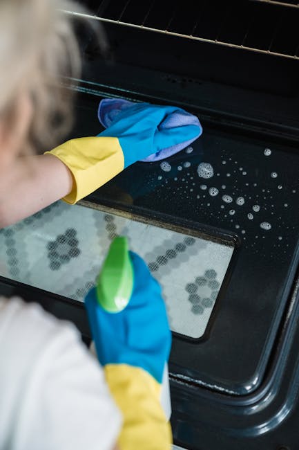 A woman wearing yellow and blue cleaning gloves is seen deep cleaning the interior of a black oven in a modern kitchen. She is holding a spray bottle in one hand and a cleaning cloth in the other, met