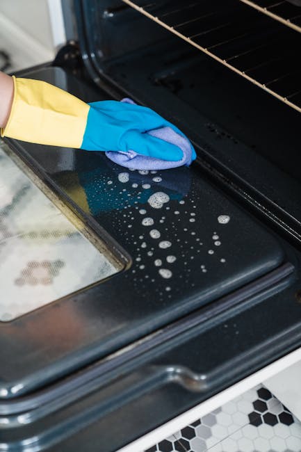 Close-up of a person wearing a yellow and blue rubber glove, cleaning the black glass surface of an oven door with a soapy blue cloth. The oven door is partially open, revealing the interior with a metal rack. The surrounding areas of the oven show a clean and shiny surface, indicating thorough surface cleaning as part of a deep cleaning process. The floor visible at the bottom features black and white hexagonal tiles, and natural light illuminates the scene, highlighting the cleanliness. This image exemplifies professional domestic cleaning practices, as performed by Oven Cleaning Merton, highlighting the importance of detailed surface sanitation in oven maintenance to prevent grease build-up risks, as discussed on the 'Grease build-up risks: emergency oven cleaning in Colliers Wood, Merton' page.
