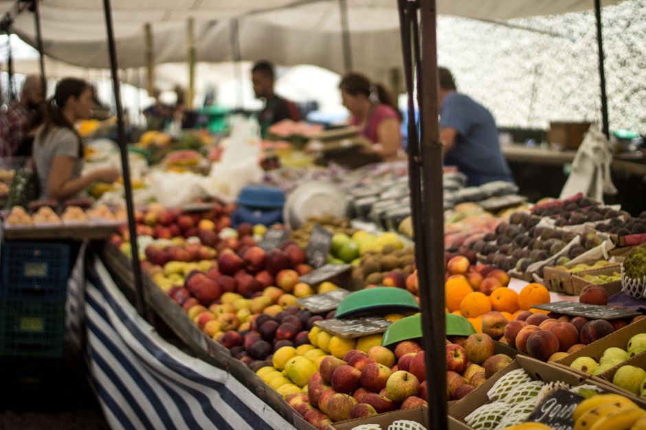 A woman wearing a gray, padded winter coat and dark pants stands at an outdoor fruit market stall near Wimbledon Common, inspecting fresh produce. The stall features a variety of fruits including pine