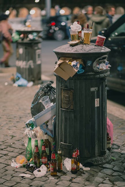 A hand wearing a yellow rubber glove reaches into an open cardboard box containing various batteries of different sizes and types, including AA and AAA, on a light-colored surface. The box is set against a neutral background, and the batteries appear to be for disposal or recycling. This scene reflects the importance of proper waste segregation and safe disposal practices in accordance with Merton Council oven waste rules, as part of domestic cleaning and waste management, in line with the guidelines highlighted on page 'Merton Council oven waste rules: safe disposal in CR4, Merton' and supported by Oven Cleaning Merton's professional cleaning services.