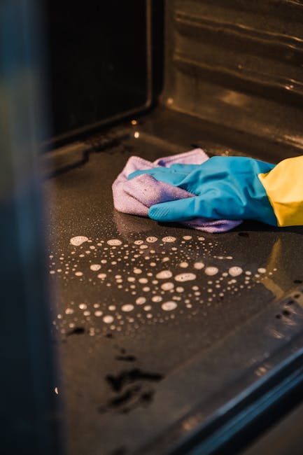 Close-up view of a black stove surface being cleaned inside a kitchen, with a gloved hand using a purple cloth to wipe away residue. The surface shows soap bubbles and streaks, indicating a recent cleaning process. The surrounding area includes stainless steel and ceramic components, with a soft light illuminating the area. The image emphasizes surface cleaning and deep cleaning techniques for sanitisation, consistent with professional domestic cleaning standards. The presence of detergent bubbles and wiping action suggests a focus on maintaining hygiene and cleanliness in kitchen appliances, aligned with oven cleaning services provided by Oven Cleaning Merton.