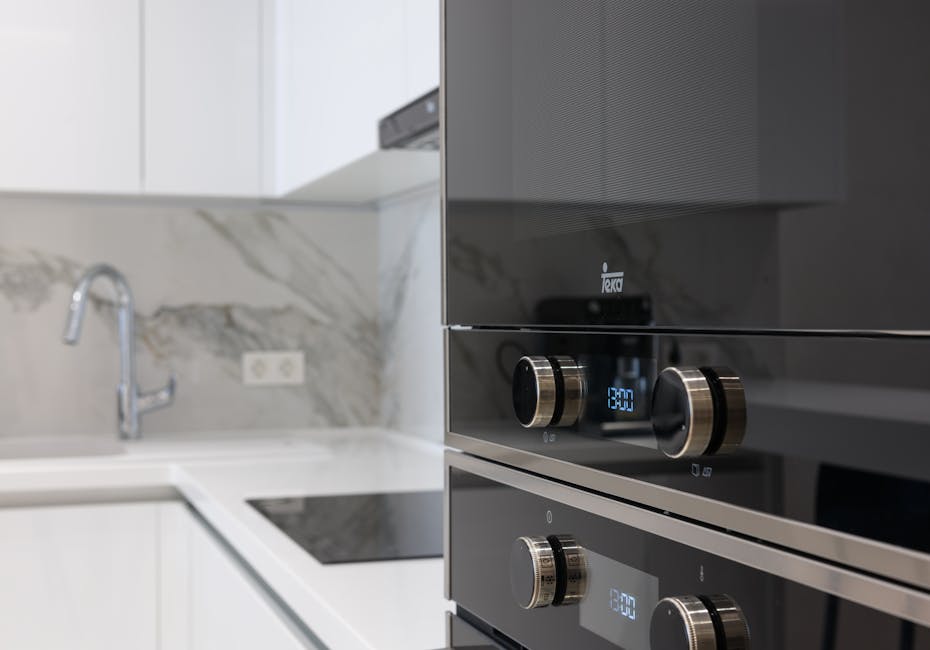 Close-up of a person's hand wearing a blue glove and a yellow sleeve, applying a cleaning spray to the black glass surface of an oven door. The oven is open, revealing stainless steel racks inside, and small soap bubbles are visible on the surface indicating active cleaning. The surrounding countertop is visible with a patterned black-and-white tiled floor beneath, suggesting a modern kitchen setting. The image emphasizes surface cleaning and deep sanitisation processes, exemplifying professional oven cleaning services offered by Oven Cleaning Merton, as referenced in the page titled 'Oven cleaning costs in Merton: what a quote should include' on ovencleaningmerton.com.