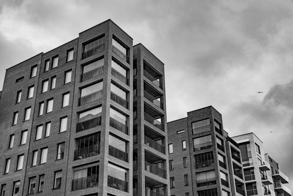 A black-and-white photograph of a modern multi-storey residential building with multiple balconies featuring metal railings, large glass windows, and brick exterior walls. The building's façade displays a uniform pattern of windows and balconies, with some windows reflecting light. The structure is situated against a cloudy sky, with a few birds visible in the distance, emphasizing an urban environment. The image showcases an architectural style typical of contemporary apartment blocks within the Merton area, capturing the clean lines and organized layout of the residential complex. While the interior cleaning services of Oven Cleaning Merton are not depicted, this image aligns with the themes of property maintenance and hygiene relevant to the PAGE_TITLE, emphasizing the importance of clean, well-maintained living spaces in flats and blocks managed or cleaned by [COMPANY_NAME].