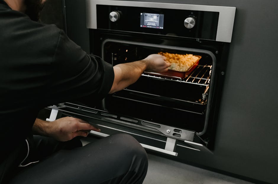A person wearing a black shirt is placing a tray of cooked rice into a modern stainless steel oven with a digital display and control knobs. The oven's interior is illuminated, showing clean glass and metal surfaces. The surrounding kitchen wall is painted dark grey, and the oven is built into a cabinet. The individual is kneeling or sitting on the floor, focusing on surface cleaning or food placement, demonstrating domestic oven use and maintenance. The scene highlights the cleanliness and functional state of the oven, typical of professional cleaning services provided by Oven Cleaning Merton.