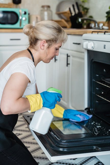 A woman wearing a white t-shirt and yellow and blue rubber gloves is seen cleaning the interior of a black glass oven door with a spray bottle and a blue cloth in a modern kitchen. The oven, with a stainless steel handle, is part of a white kitchen unit with black handles, surrounded by various kitchen appliances and utensils on wooden countertops, including a teal microwave and glass jars. The scene is well-lit with natural light, highlighting the cleanliness of the surfaces, including the shiny oven and the surrounding ceramic tiles and wooden worktop, demonstrating thorough surface cleaning and sanitisation. The setting emphasizes domestic cleaning and deep cleaning processes, with a focus on hygiene and maintenance by Oven Cleaning Merton, relevant to the service offered on the webpage titled 'Same-day oven cleaning in Wimbledon SW19 - prices, Merton.'